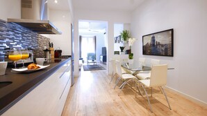 A modern kitchen with a breakfast bar, a dining area with a glass table and chairs, and a cityscape painting on the wall.