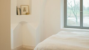A bedroom with a large bed, a window, and a wall-mounted shelf with books and a framed picture. - Apartment Gut Üselitz on Rügen - 1. First floor left (Poseritz)
