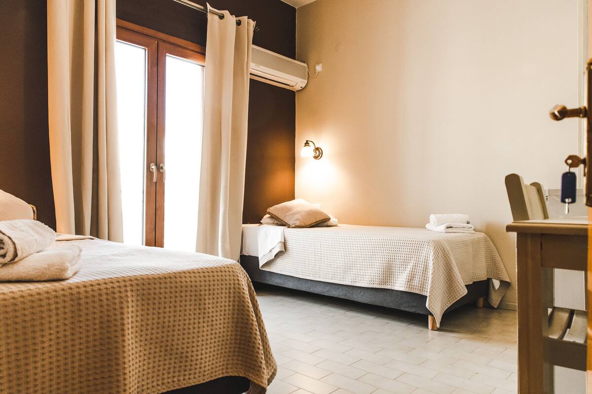 A hotel room with two beds, a window with curtains, a wall-mounted air conditioner, and a bedside table with a lamp.