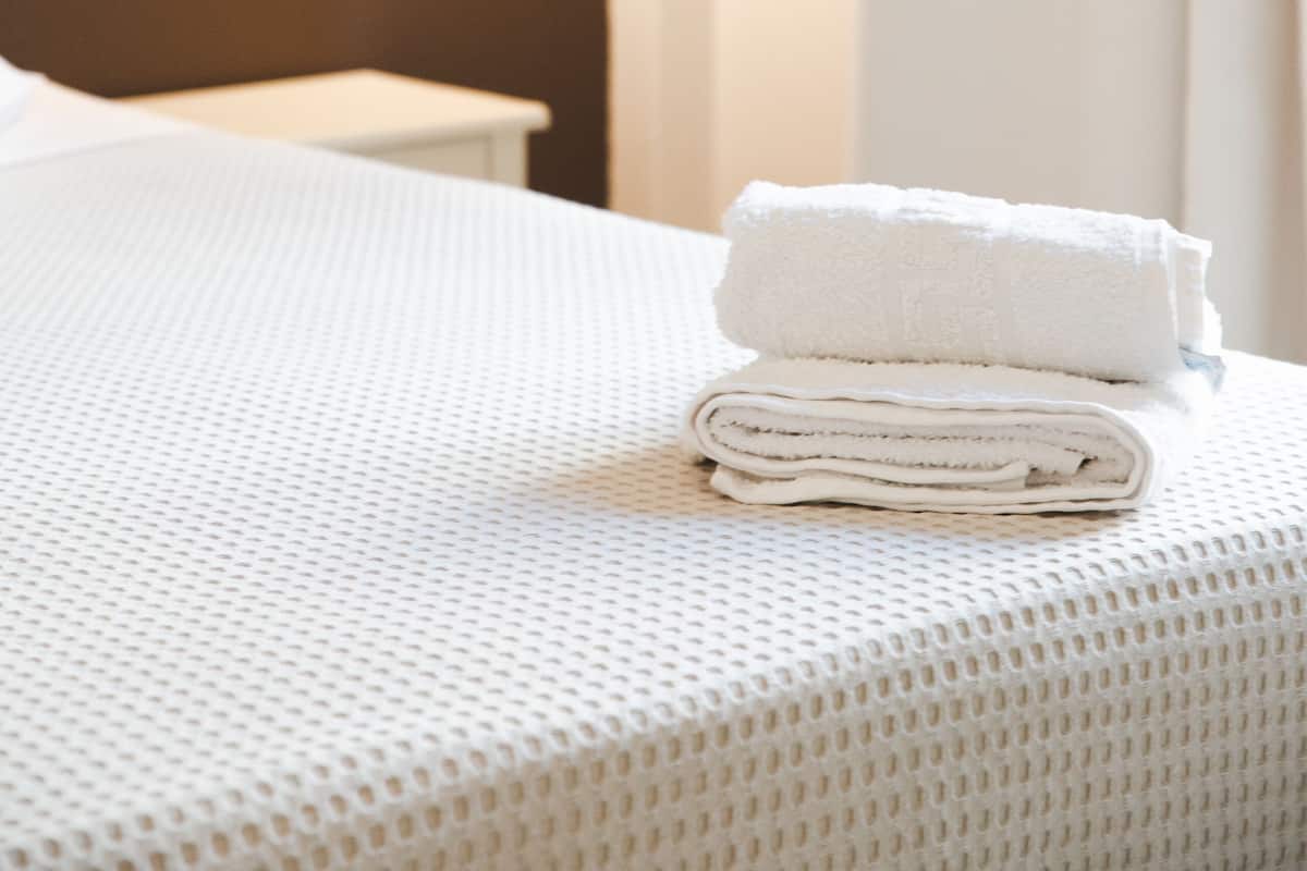A neatly made bed with a folded white towel, a wall-mounted light fixture, and a plain wall.