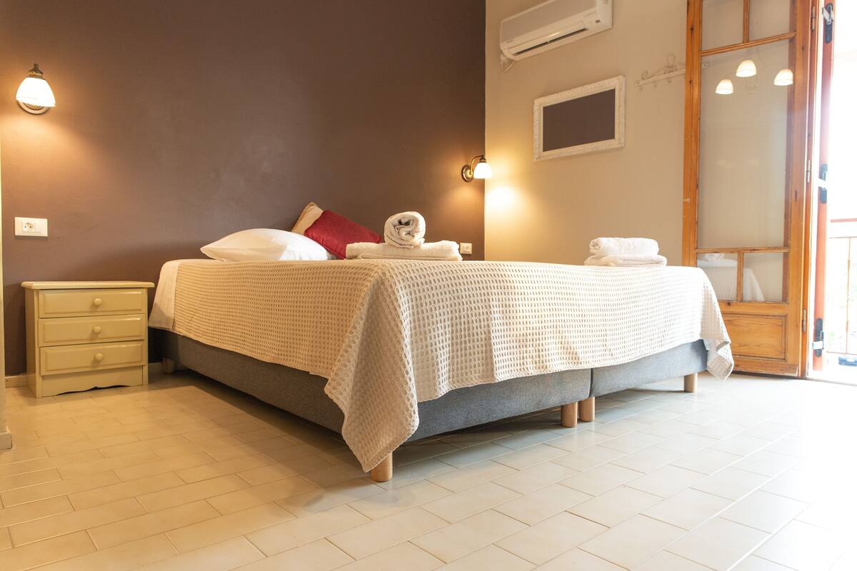 A neatly made bed with a white blanket and two towels, a nightstand with drawers, a wall-mounted light, and a window with blinds.