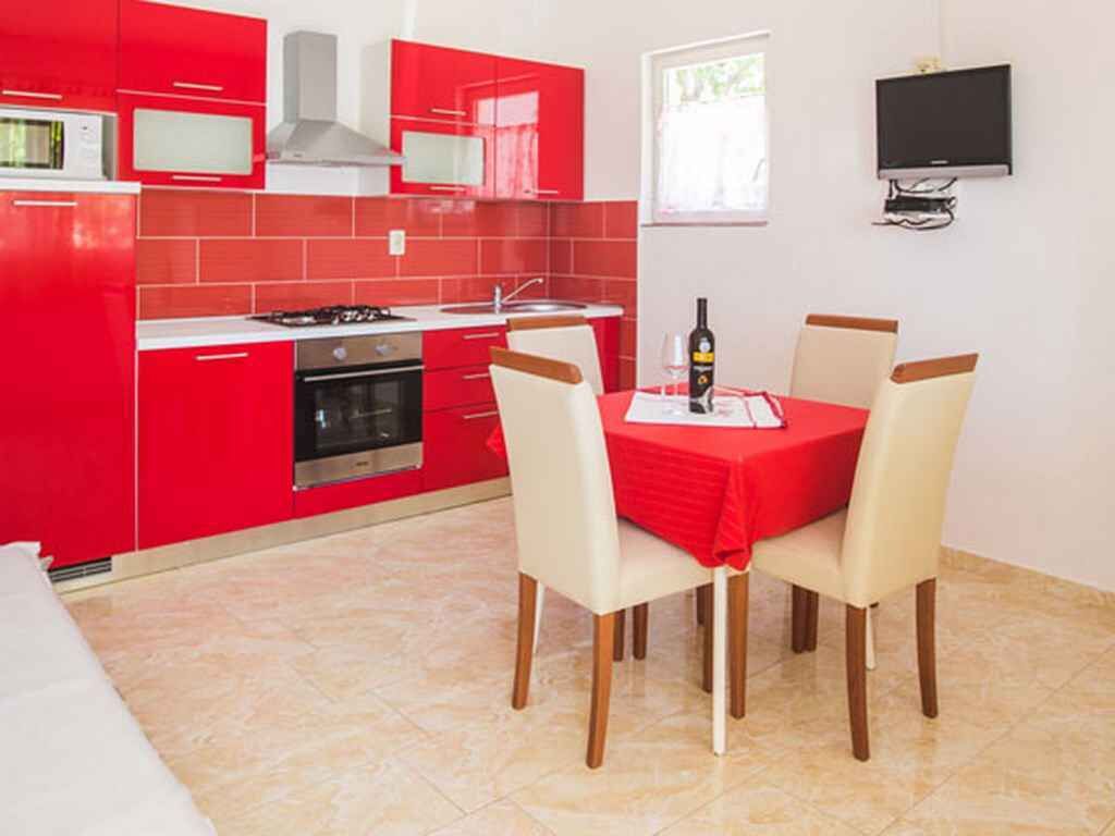 A compact kitchen with red cabinets, a gas stove, and a microwave. There is a dining table with four chairs and a TV mounted on the wall.