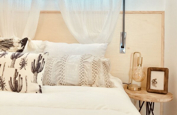 Una cama bien hecha con un cojín blanco y estampado, una lámpara de noche y una mesita de noche de madera con un marco de fotos.