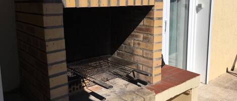 A brick-built outdoor grill with a metal grate, positioned against a wall with a sliding glass door.