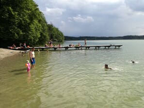Escena junto al lago con un muelle que se adentra en el agua, rodeado de vegetación y gente disfrutando del entorno.
