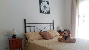 A neatly made bed with patterned bedding, a bedside lamp, a small bedside table, a teddy bear, and a wall-mounted flower painting.