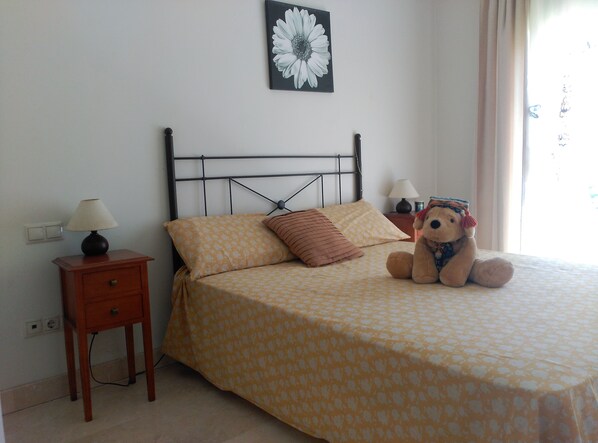 Un lit bien fait, avec une literie à motifs, une lampe de chevet, une petite table de chevet, un ours en peluche et un tableau représentant des fleurs accroché au mur.