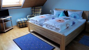 Un dormitorio con techo inclinado, una cama con ropa de cama floral, una silla, un televisor sobre un soporte y una mesita.