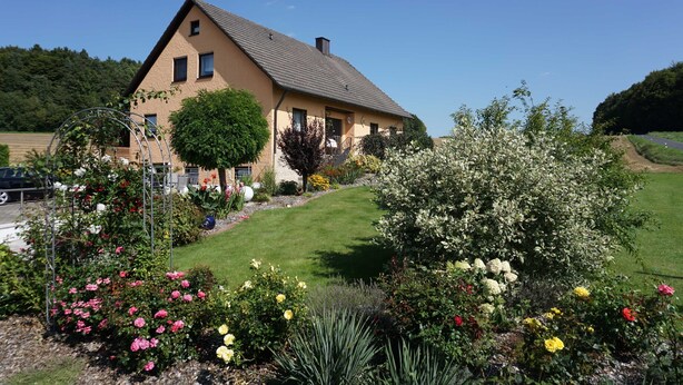 Una casa de dos pisos con techo inclinado, rodeada por un jardín bien cuidado con diversas flores y arbustos.