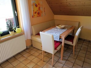 Una habitación acogedora con una mesa de madera, sillas blancas, un sofá, un radiador, una planta en maceta y una ventana con cortina.