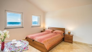 Une chambre à coucher comprenant un lit, des tables de chevet, une petite table avec un vase et des tasses, et deux fenêtres donnant sur la mer.