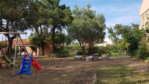 Un terrain de jeux avec un toboggan et des balançoires, entouré d’arbres et bordé d’un bâtiment surmonté d’un toit en forme de dôme.