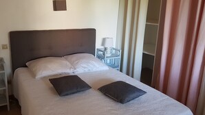 Un lit bien fait, avec deux oreillers et deux couvertures pliées, une table de chevet avec une lampe, et une vue sur un placard aux portes coulissantes.