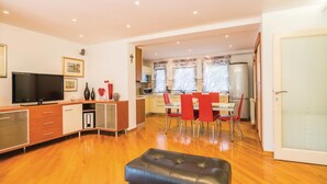 Een moderne woonkamer met een flatscreen televisie, houten vloeren, een eethoek met rode stoelen en een keuken met een koelkast en magnetron.