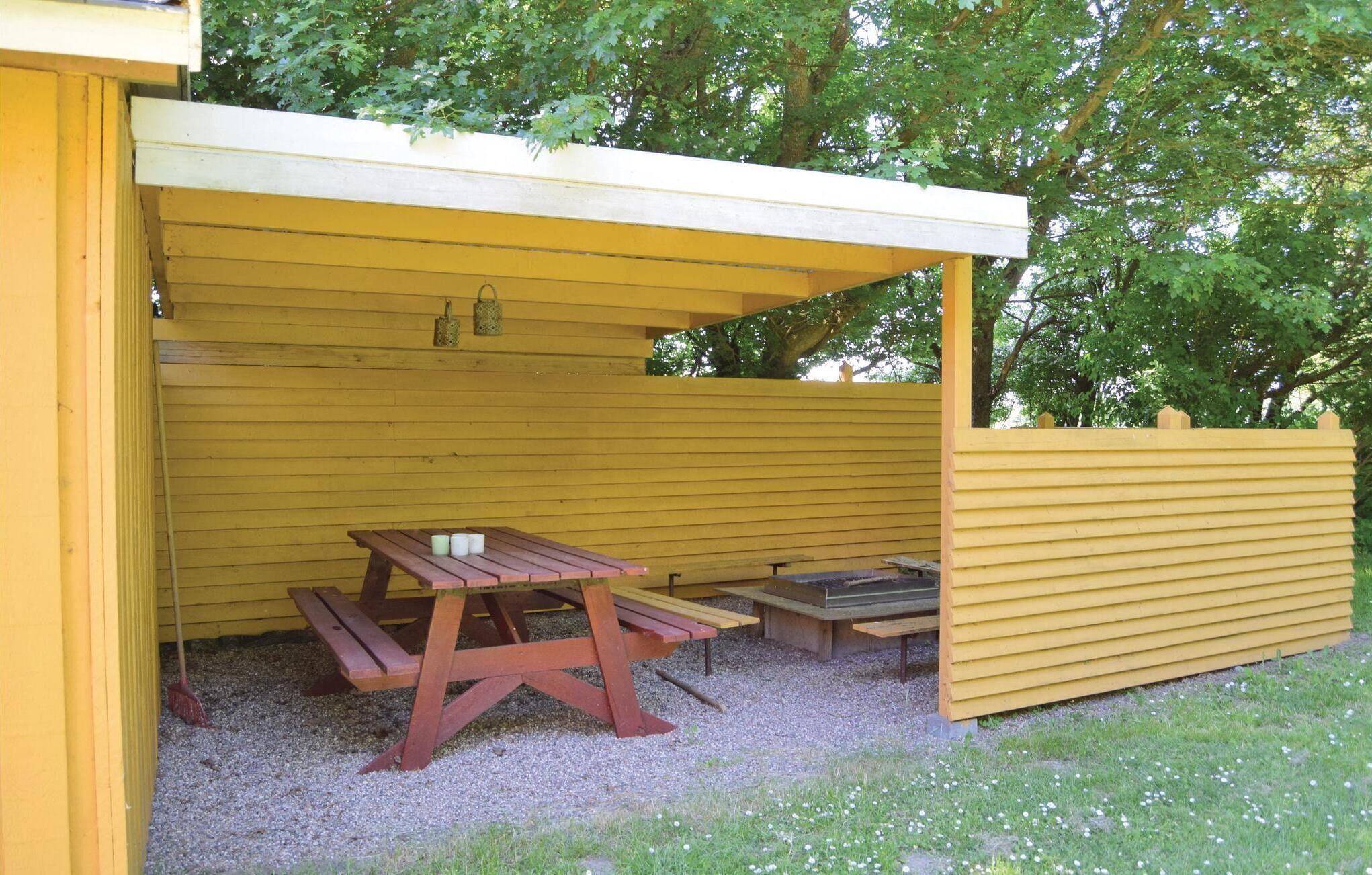 Ein überdachter Picknickbereich mit Bänken und Tisch, umgeben von einem gelben Zaun und Bäumen.