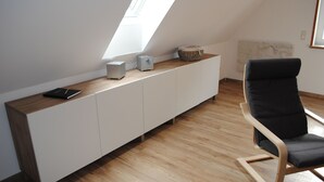 Une pièce moderne avec un plafond en pente, un meuble blanc sur lequel est posée une tablette, une chaise noire et un petit haut-parleur.