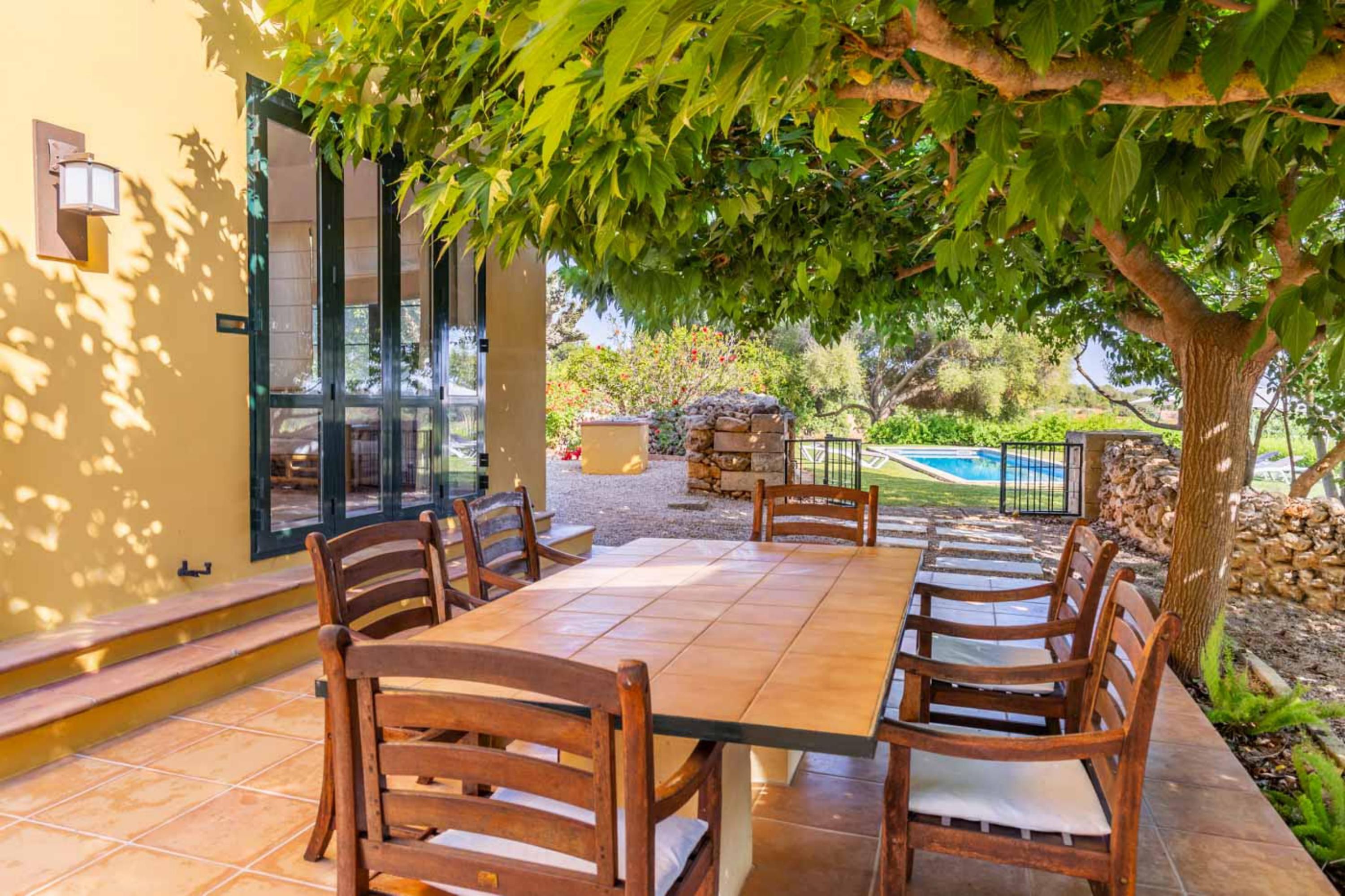 Une terrasse avec une table et des chaises en bois, entourée d’un mur de pierre et d’un arbre qui offre de l’ombre.