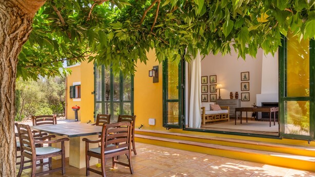 A dining area with a table and chairs, a window with a view of the interior, and a tree providing shade. - 784524 Ses Cases, Spain (Manacor)