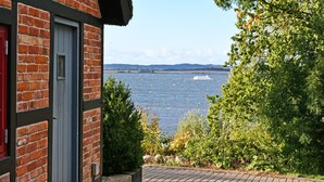 A brick building with a red door, a view of a body of water, and greenery in the foreground. - 01 Holiday apartment Gänseblümchen with lake view - Haus am Höft in Gager with lake view (Gager)