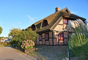 A traditional half-timbered house with a thatched roof, a cobblestone driveway, and a flower bed with pink flowers.