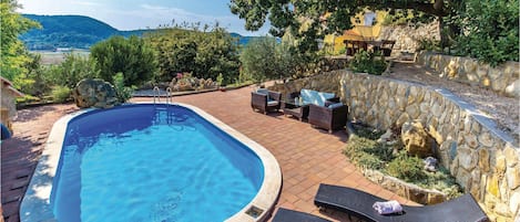 A swimming pool with lounge chairs and a stone wall, surrounded by greenery and a view of hills.