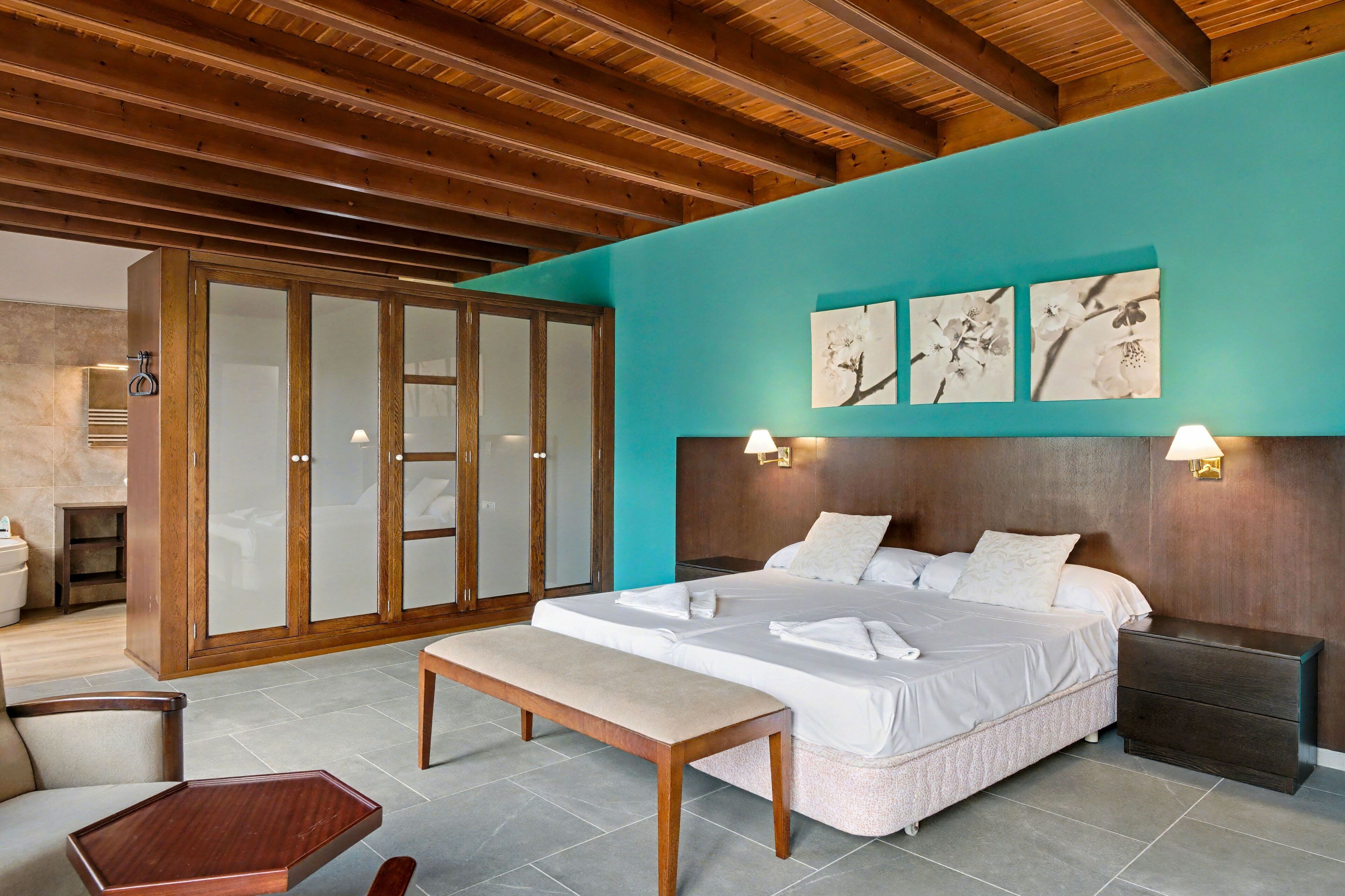 Une chambre avec un plafond en bois, un mur d’accent de couleur turquoise, un lit avec du linge de lit blanc, un banc et une table de chevet.