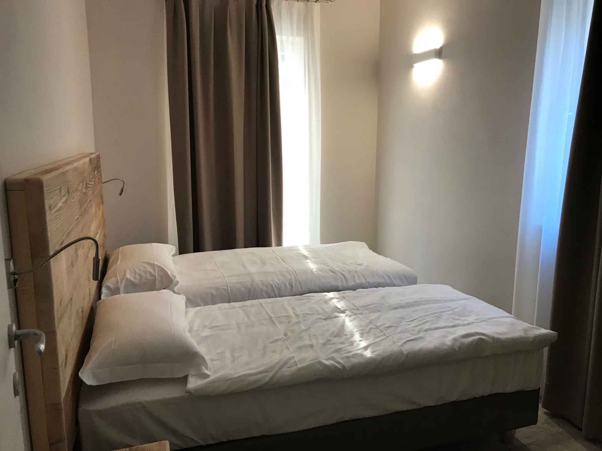 Een net opgemaakt bed met witte lakens in een kamer met een houten hoofdeinde, een raam met gordijnen en wandlampen.