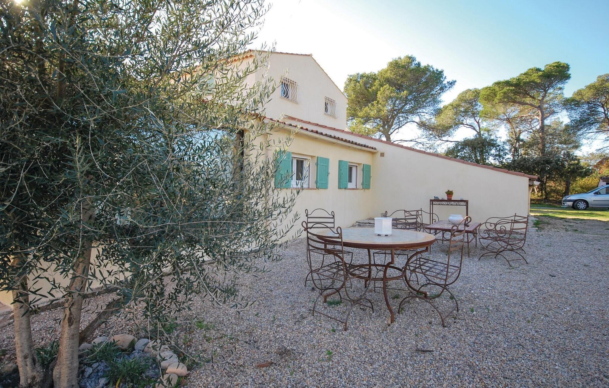 A rustic outdoor seating area with metal chairs and a table, surrounded by olive trees and a building with green shutters.