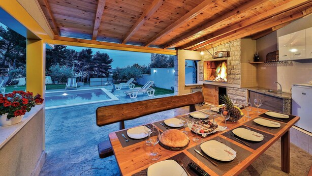 Un área de comedor exterior acogedora, con una mesa de madera preparada para una comida, una chimenea de piedra y vistas a una piscina y jardín.