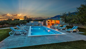 Una piscina con sillones de descanso, un edificio de piedra y vista al atardecer.