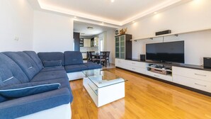 Una sala de estar moderna con un sofá seccional, una mesa de centro de vidrio, un televisor de pantalla plana y una zona de cocina al fondo.