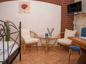 Una habitación con una cama, dos sillones de mimbre, una mesita con una lámpara y un televisor en la pared.
