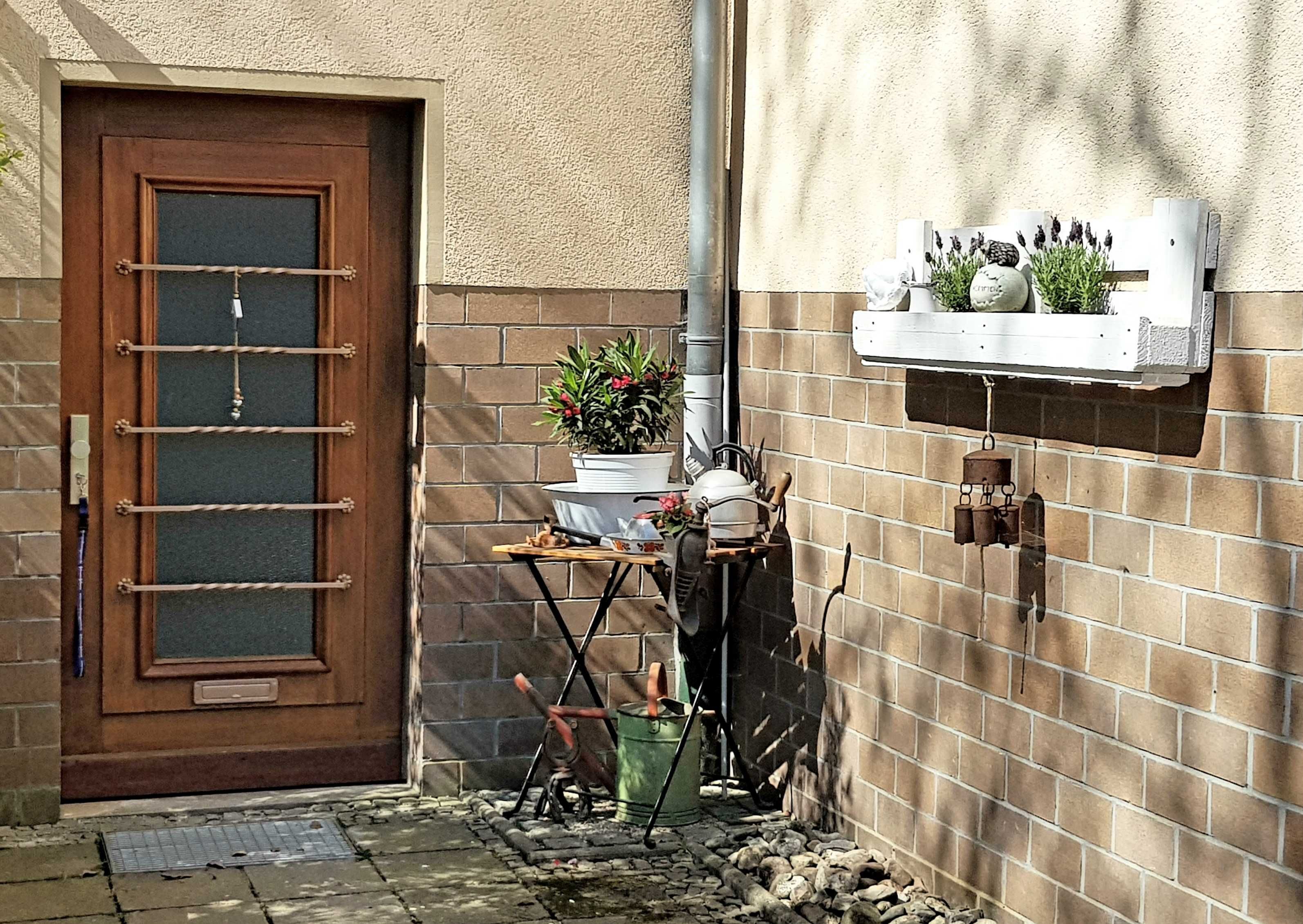 Een deur met een ruit en een metalen hekwerk, een kleine tafel met tuingereedschap, een potplant en een wandplank met decoratieve objecten.