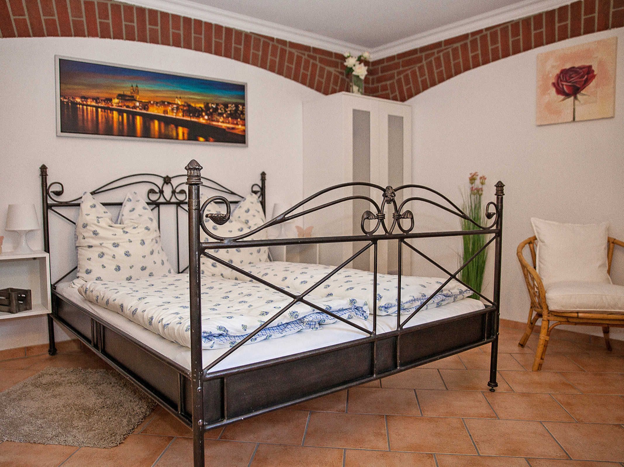 A room with a metal-framed bed, a white chair, a painting of a cityscape, and a vase with flowers.