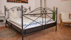 A room with a metal-framed bed, a white chair, a painting of a cityscape, and a vase with flowers. - Apartment Sandra Mattaj (Blankenburg (Harz))