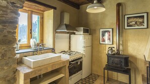 Una cocina rústica con pared de piedra, vigas de madera, un lavamanos blanco, estufa, refrigerador y una pequeña mesa negra con una tetera.