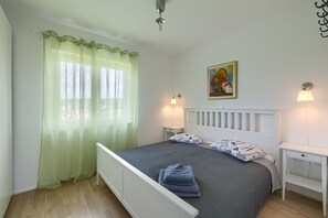 A bedroom with a bed, two bedside tables, a nightstand, a lamp, a painting, and a window with sheer curtains. - Casa Leone (Sutivanac)