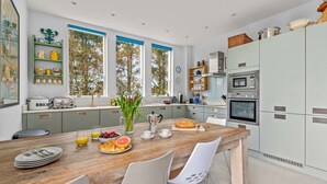 Une cuisine moderne dotée d’un îlot central en bois, d’appareils électroménagers intégrés et offrant une vue sur les arbres grâce à de grandes fenêtres.