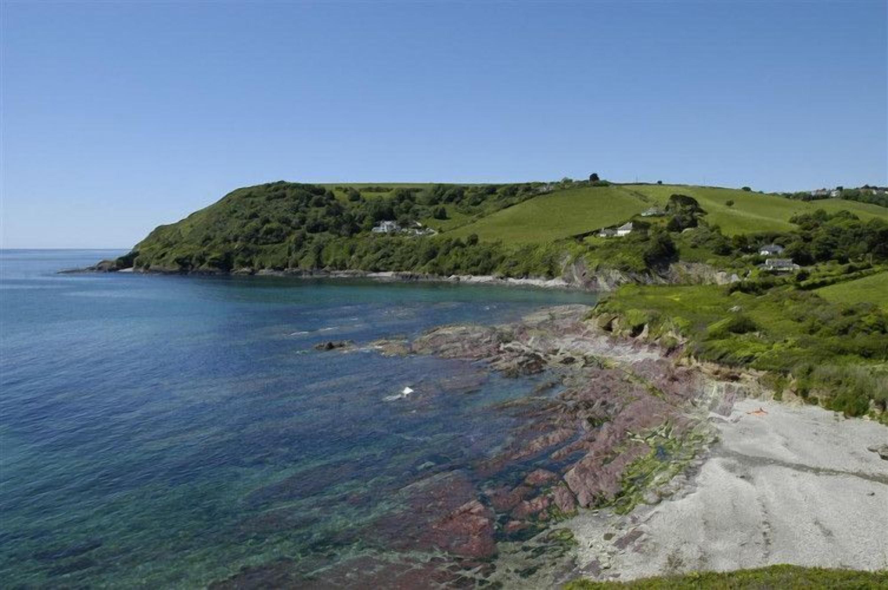 A coastal landscape with a clear view of the sea, green hills, and a few scattered houses.