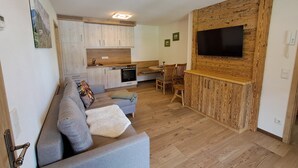 A modern living room with a grey sofa, wooden flooring, a kitchen with light wood cabinets, a dining area, and a mounted TV. - Vacation apartment 4 Resi - Guesthouse Becher, Kreuth-Point (Kreuth)