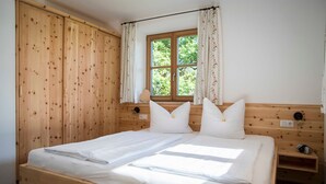 A wooden bed with white linens, a window with curtains, and a small bedside table. - Vacation apartment 4 Resi - Guesthouse Becher, Kreuth-Point (Kreuth)