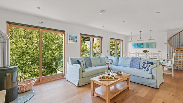 A spacious living room with a light blue sofa, a wooden coffee table, and large windows offering a view of greenery. - Beautifully decorated "Sea Shack", with large sun roof terrace and large private hot tub. (Looe)