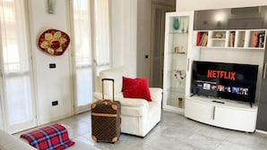 Ein Wohnzimmer mit einem weißen Sofa, einem roten Kissen, einem braunen Koffer, einem Netflix-Fernseher und einer Vitrine mit Büchern.