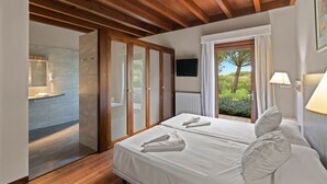 A hotel room with two beds, a wooden ceiling, a view of greenery, a bathroom with a shower, and a TV on the wall.