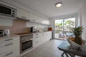 A modern kitchen with white cabinets, a microwave, and a coffee maker. There is a dining table with a fruit bowl containing a pineapple and oranges.