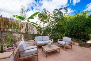 An outdoor patio area with a wooden table and two sofas, surrounded by plants and a bamboo fence.