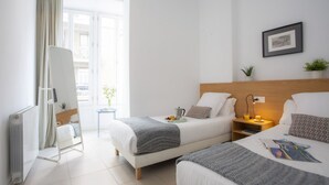A hotel room with two beds, a bedside table with a lamp, a small table with a plant, and a window with curtains. - Singular Stays Mar44 VII (Valencia)