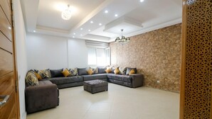 A spacious living room with a large L-shaped sectional sofa, a small ottoman, and a decorative wall with a stone pattern. - Appartement 26 ensoleillé à 5 min de la plage El Jadida (El Jadida)