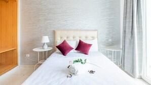 A neatly made bed with a white duvet, two purple pillows, and a rolled towel with a green leaf. There is a small white table with a lamp beside the bed. - Appartement 26 ensoleillé à 5 min de la plage El Jadida (El Jadida)