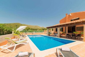 A swimming pool with lounge chairs, a patio area with tables and chairs, and a view of hills in the background.